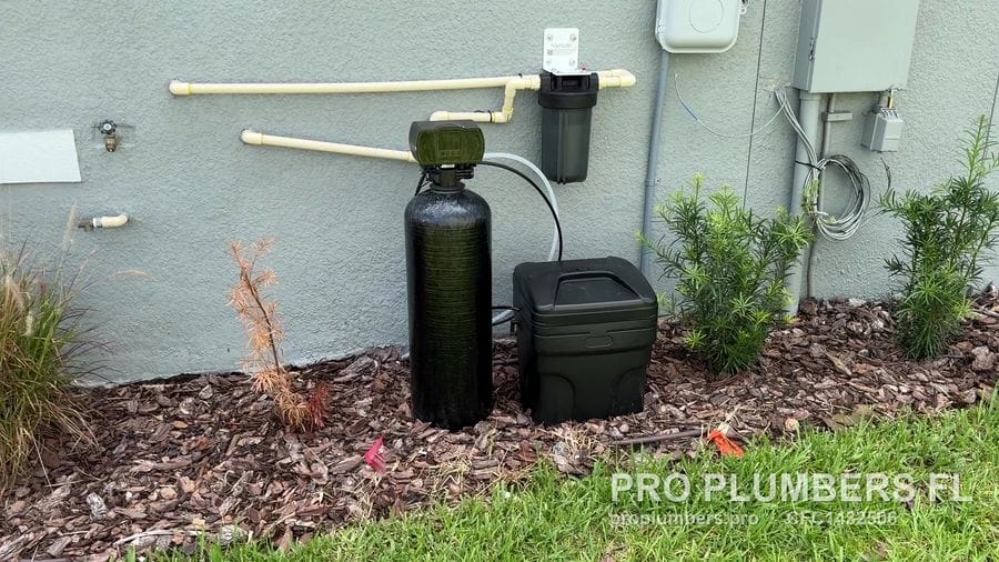 Exterior water softener, brine tank and prefilter on a mulch bed against a stucco wall in Wesley Chapel, Florida by PRO PLUMBERS FL, LLC (licence CFC1432506)