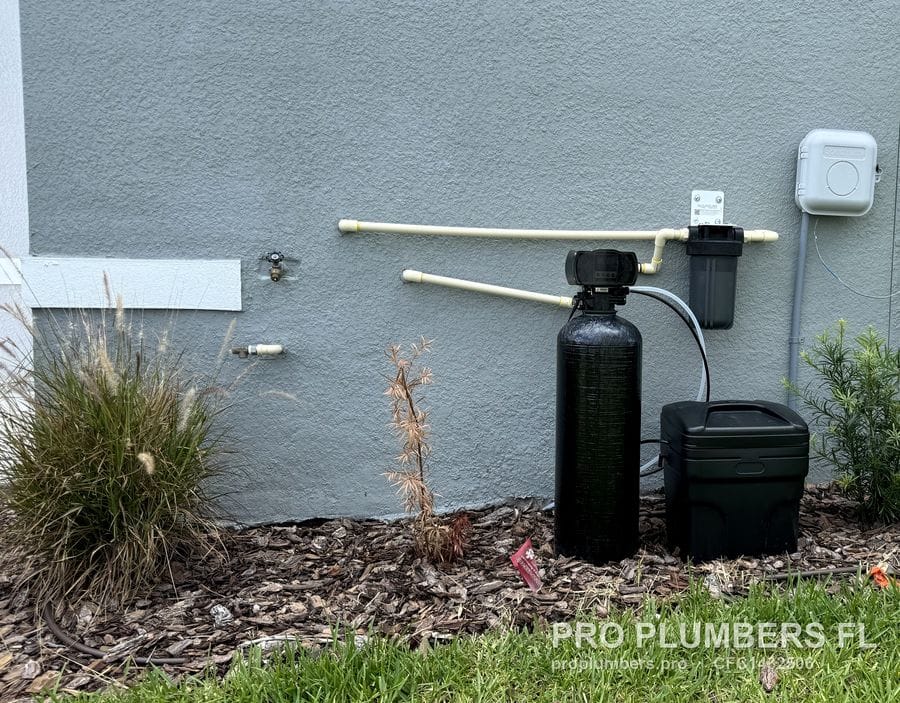 Exterior water softener, brine tank and prefilter installed along a grey stucco wall on a mulch bed in Wesley Chapel, Florida by PRO PLUMBERS FL, LLC (licence CFC1432506)