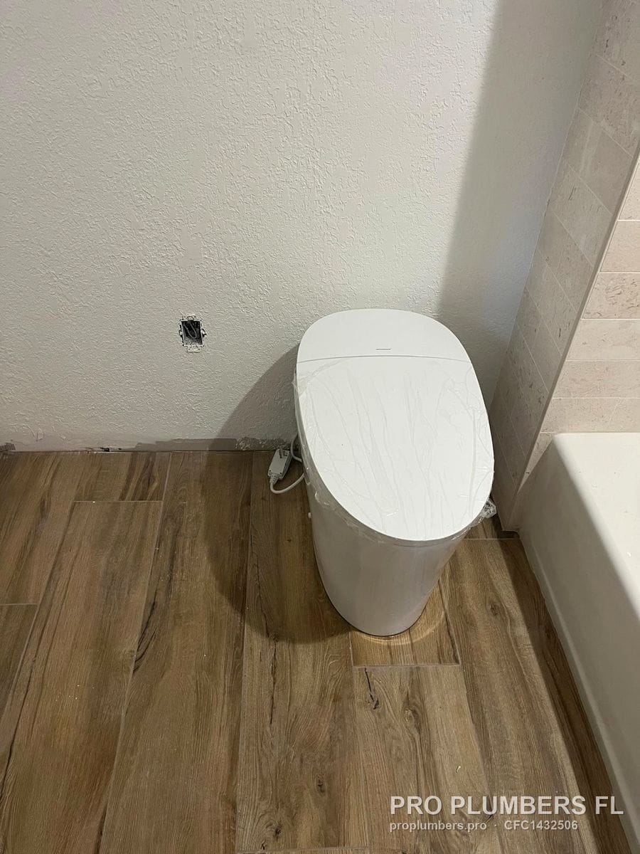 Smart toilet in protective film installed on wood-look tile with power outlet box above in Clearwater, Florida by PRO PLUMBERS FL, LLC (licence CFC1432506)