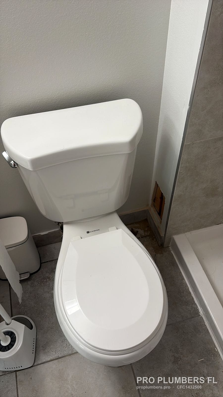 Two-piece round-front toilet with new seat on grey floor tile next to a shower base in Clearwater, Florida by PRO PLUMBERS FL, LLC (licence CFC1432506)