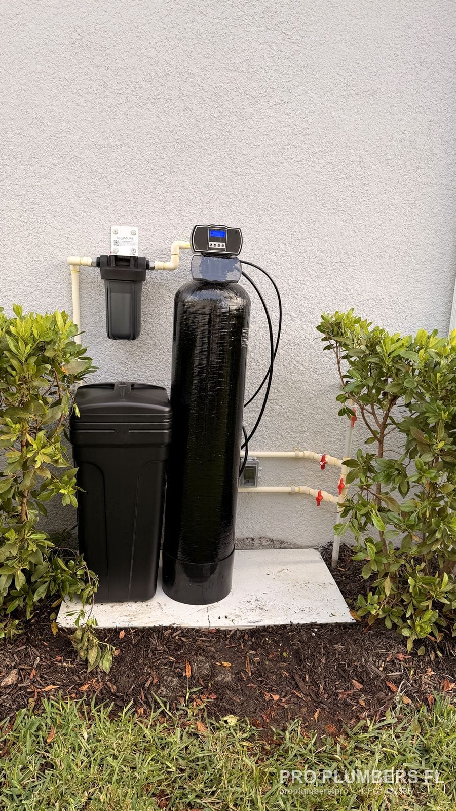 Water softener and brine tank on a concrete pad with wall prefilter and CPVC bypass valves between shrubs in Sarasota, Florida by PRO PLUMBERS FL, LLC (licence CFC1432506)