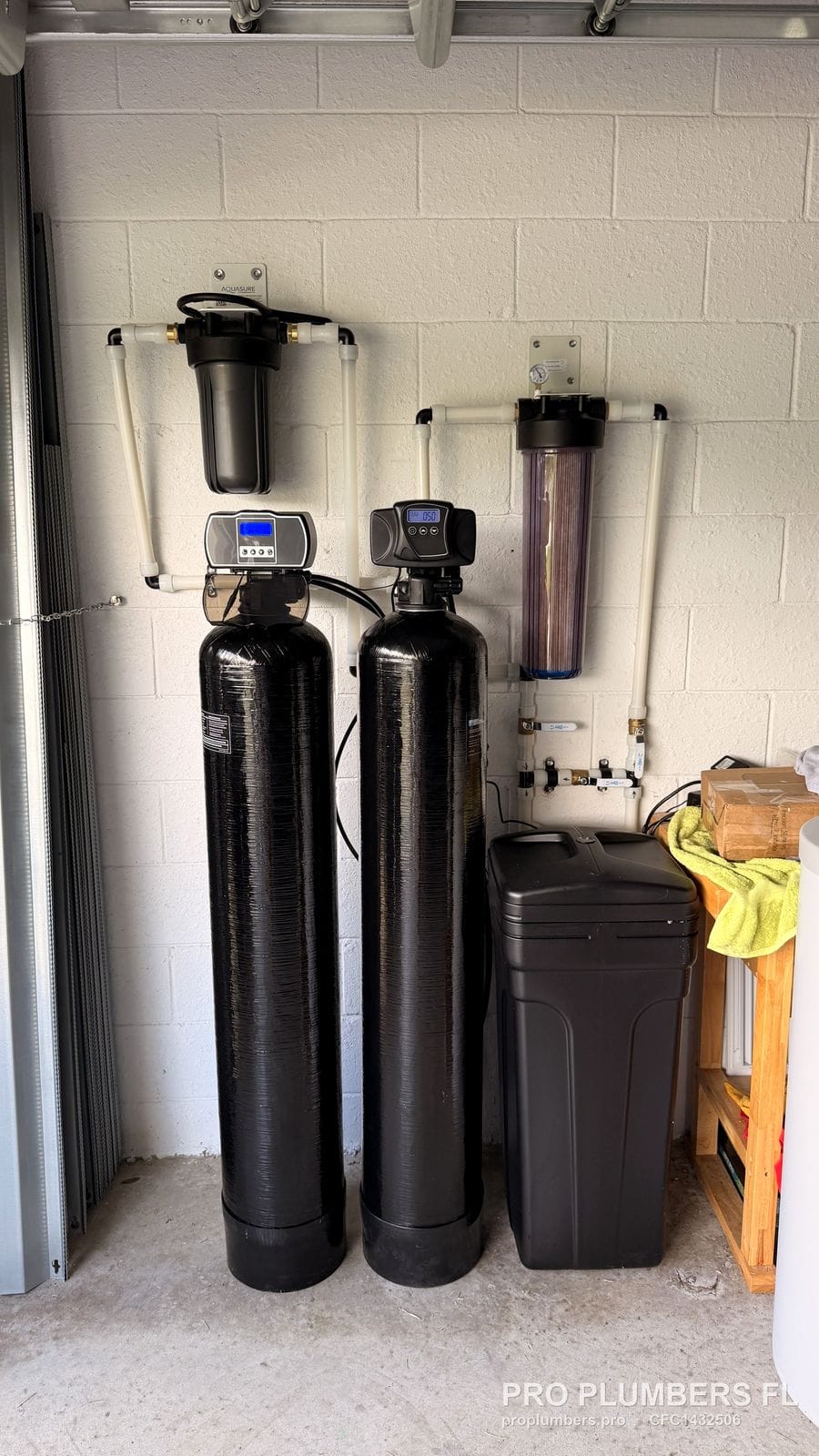Full-height view of a two-tank softener and filter system with brine tank on a garage wall in Bradenton, Florida by PRO PLUMBERS FL, LLC (licence CFC1432506)